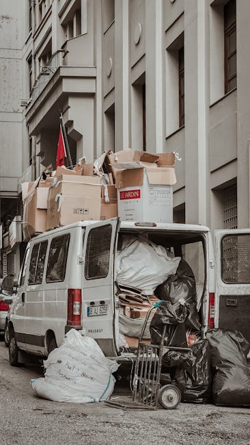 A white moving van parked on a city street next to a multi-story residential building, with its rear doors open revealing an interior filled with various packed items and furniture. On top of the van, several large cardboard boxes and a wrapped mattress are stacked, indicating a home relocation process. Outside the van, additional black and white rubbish bags, packing materials, and a small hand trolley are visible on the pavement, suggesting the loading or unloading of belongings. The building features tall windows and typical urban architecture, with a red flag attached to the wall nearby. The scene captures the logistics of furniture transport and packing during a house removal in Notting Hill, facilitated by Man with Van Notting Hill, highlighting the practical steps of loading used during same-day removal services.