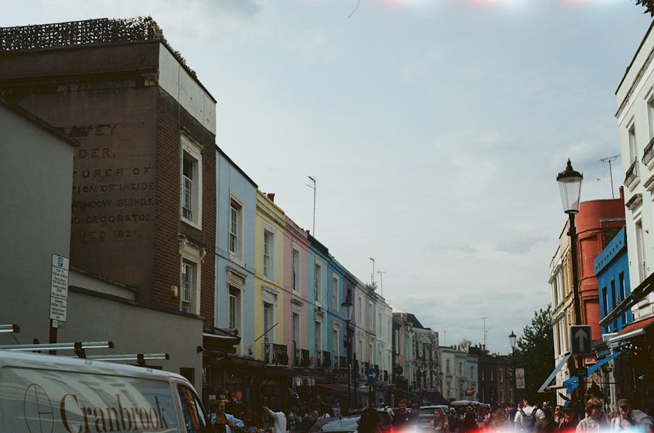 A vintage red and white van parked on the street in front of pastel-colored Victorian-style houses in Notting Hill, with a large leafless tree in the foreground. The van is equipped with a bike rack o