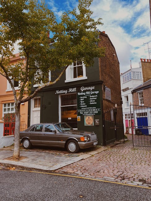 The exterior of a dark green, two-story building with a sign reading 'Notting Hill Garage' and listing services such as removals, storage, and packing supplies. A silver car is parked on the cobblestone pavement directly in front of the garage entrance. A medium-sized tree with green leaves partially obscures the upper part of the building. The surroundings include neighboring residential buildings with white and brick facades, and pathways leading to the garage entrance. The scene is lit by daylight with some clouds in the sky, indicating an overcast day. This setting depicts a typical home relocation environment, with the garage serving as a base for furniture transport and packing during moving services, as provided by Man with Van Notting Hill.