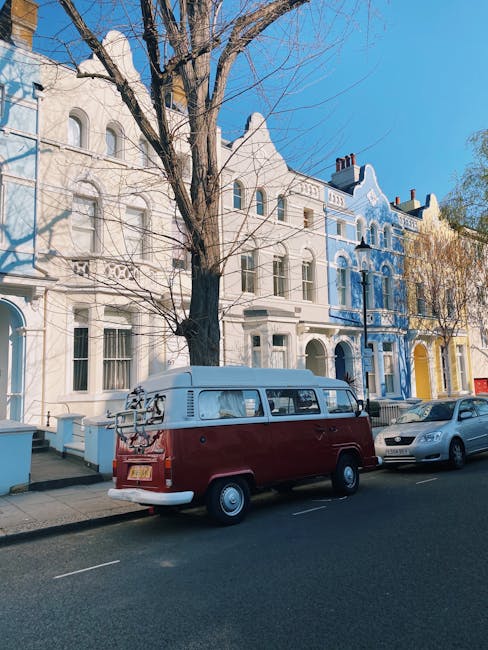 Image showing a row of Victorian-style terraced houses, with the first building painted in bright red and the next in pale blue. The red house has a small front yard with concrete steps leading up to a door, flanked by two white rectangular planters containing green shrubs. The steps are partially covered with a red-painted ramp that appears to be used for moving furniture or other large items. A black iron fence runs along the sidewalk in front of the houses, with a brick pillar supporting it near the red house. The scene is well-lit with natural daylight, and the sky is clear and blue, indicating a sunny day. This setting exemplifies typical London residential architecture and highlights the exterior environment involved in home relocation and furniture transport services, as offered by Man with Van Notting Hill.