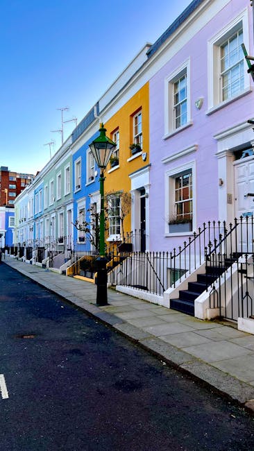 A row of colorful terraced houses on a quiet residential street with a black iron fence and steps leading up to each entrance. The houses are painted in pastel shades of blue, yellow, purple, and pink, with large sash windows and white trims. A vintage-style street lamp with a green top and black base stands on the pavement in front of the houses. The street has a paved sidewalk with some small leafless trees planted along the edge, and the sky above is clear and blue. This scene depicts a typical London neighbourhood, suitable for home relocation or furniture transport in the context of house removals, as part of a professional moving process by Man with Van Notting Hill.