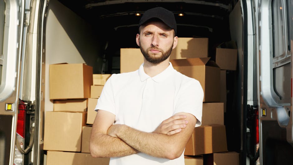 A man dressed in a blue hoodie and matching trousers is standing at the open back of a white delivery van parked on a cobblestone street in an urban residential area. He is holding a small cardboard b