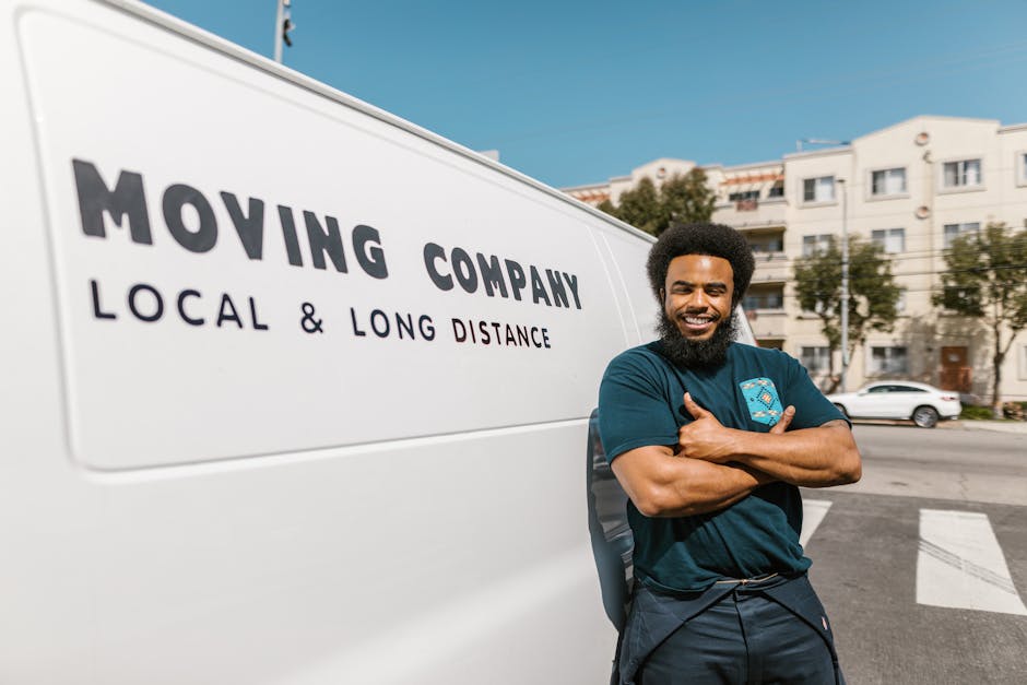 A smiling male mover with dark curly hair and a beard, wearing a navy blue T-shirt, stands with arms crossed next to a white moving van that has the text 'MOVING COMPANY LOCAL & LONG DISTANCE' printed on its side. The van is parked on a street in a residential area, with white apartment buildings and a few cars visible in the background. Inside the open doorway of the van, various furniture items and moving boxes are visible, including cardboard boxes wrapped in plastic and blankets. The interior of the van shows a loading process for a home relocation, with items arranged inside in preparation for moving. The scene is captured under clear daylight with a blue sky, reflecting a professional and organized house removal service by Man with Van Notting Hill, assisting with furniture transport and packing during a house move in W11.