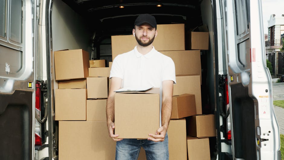 A man wearing a black cap, white polo shirt, and blue jeans stands inside the open rear doors of a moving van, holding a cardboard box in front of him. The van's interior is filled with neatly stacked cardboard boxes of various sizes, some on the floor and others arranged on shelves or stacked against the sides. The boxes are sealed with packing tape, and a few are wrapped in plastic or fabric covers. The outside environment shows a residential street with houses and a pavement, indicating a home relocation process. The image captures the loading or unloading phase of furniture transport, possibly part of a house removal service by Man with Van Notting Hill, designed for efficient packing and logistics during a move in W11.