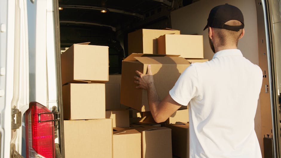A smiling male mover with dark curly hair and a beard, wearing a navy blue T-shirt, stands with arms crossed next to a white moving van that has the text 'MOVING COMPANY LOCAL & LONG DISTANCE' printed on its side. The van is parked on a street in a residential area, with white apartment buildings and a few cars visible in the background. Inside the open doorway of the van, various furniture items and moving boxes are visible, including cardboard boxes wrapped in plastic and blankets. The interior of the van shows a loading process for a home relocation, with items arranged inside in preparation for moving. The scene is captured under clear daylight with a blue sky, reflecting a professional and organized house removal service by Man with Van Notting Hill, assisting with furniture transport and packing during a house move in W11.