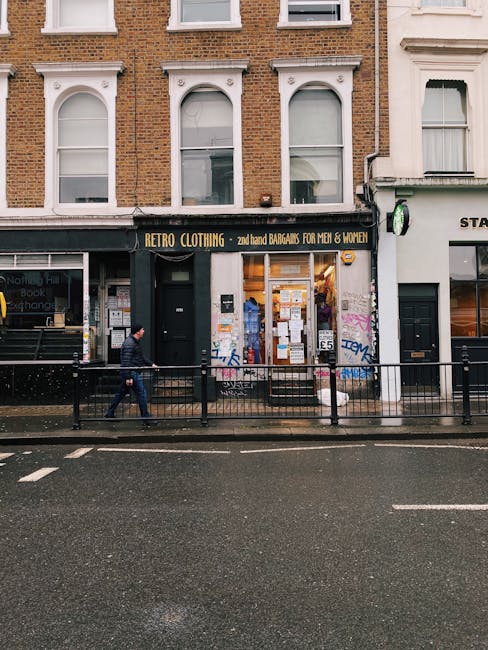The image shows the exterior of a small retail shop named 'RETRO CLOTHING,' situated on the ground floor of a multi-story building with a brick façade on the upper levels and large display windows on the shop front. The shop is located on Portobello Road, as indicated by the context of the page title, and features a black sign with yellow lettering. Outside the shop, a person dressed in dark clothing, including a black jacket and blue jeans, walks along the pavement while carrying a bag. The pavement is separated from the road by a metal railing, and the road surface appears wet, suggesting recent rain. The shop windows display various posters, notices, and merchandise, with some graffiti visible on the lower part of the wall next to the shop entrance. This scene is typical of an area involved in home or commercial property relocations, where furniture or belongings may be handled in connection with removals or packing and moving services, as provided by companies like Man with Van Notting Hill.
