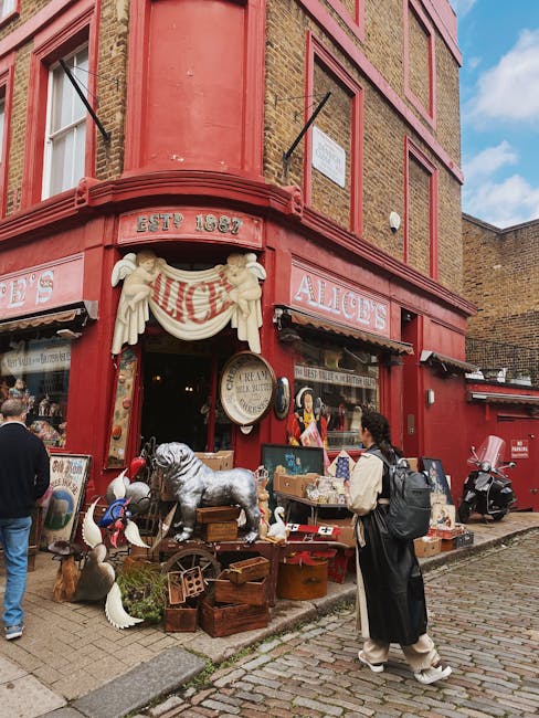 The image shows the exterior of a small retail shop named 'RETRO CLOTHING,' situated on the ground floor of a multi-story building with a brick façade on the upper levels and large display windows on the shop front. The shop is located on Portobello Road, as indicated by the context of the page title, and features a black sign with yellow lettering. Outside the shop, a person dressed in dark clothing, including a black jacket and blue jeans, walks along the pavement while carrying a bag. The pavement is separated from the road by a metal railing, and the road surface appears wet, suggesting recent rain. The shop windows display various posters, notices, and merchandise, with some graffiti visible on the lower part of the wall next to the shop entrance. This scene is typical of an area involved in home or commercial property relocations, where furniture or belongings may be handled in connection with removals or packing and moving services, as provided by companies like Man with Van Notting Hill.