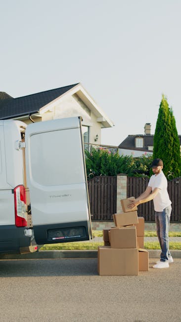 A man wearing a black cap and white polo shirt stands inside the back of a moving van, holding a cardboard box with both hands. The van's interior is filled with several stacked cardboard boxes of various sizes, some wrapped in plastic or packing materials, indicating an ongoing home relocation process. The boxes are arranged on the van's floor and shelves, with some positioned near the open rear doors. The scene is outdoors, with a residential street visible in the background, featuring houses and a sidewalk. The lighting appears natural, highlighting the details of the packing and loading process, which is typical for furniture transport and moving service activities conducted by Man with Van Notting Hill, as seen on the webpage dedicated to permits for moving vans in Notting Hill.