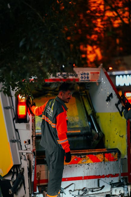 A male worker dressed in high-visibility orange and grey overalls is standing outdoors at dusk, next to a large hydraulic waste collection vehicle with reflective markings, under trees with dark foliage. The vehicle is positioned on a paved area, and the worker appears to be preparing or inspecting the equipment as part of a home relocation or waste disposal process. The vehicle's rear compartment is open, revealing a spacious interior designed for loading and transporting bulky items. The background shows a sky with a vibrant orange sunset, and ambient street lighting is visible, creating a professional and organized scene associated with Removals and logistics services suited for home removals, packing, and furniture transport within the context of safe disposal laws for mattresses and electronic waste in W11, as highlighted on the Man with Van Notting Hill website.
