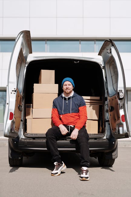 A young man with a beard, wearing a blue beanie, a navy and red hooded jacket, black pants, and white sneakers, is seated on the edge of a cargo van with its rear doors open. Inside the van, several cardboard boxes of varying sizes are arranged neatly on the floor and against the walls, awaiting transportation. The scene is set outdoors in front of a modern building with white tiled cladding and large windows, with natural daylight illuminating the setting. This image depicts the loading process involved in home relocation and furniture transport, illustrating the careful packing and preparation by [COMPANY_NAME] for a house move. The van is positioned on a paved area, and the photograph captures the final stage of loading boxes, signifying the logistics of packing and moving tasks associated with professional removals services, highlighting the importance of secure packaging and transport for household goods.