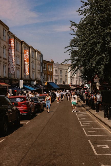 The image shows a busy street scene in Notting Hill during daytime, featuring a row of multi-storey buildings with retail storefronts and large vertical banners hanging on the facades. The street is filled with pedestrians, some walking along the pavement and others gathering outside shops. Parked cars line the sides of the road, including a prominent red vehicle near the foreground. On the right side, there is a designated parking area with marked spaces, separated from the sidewalk by black bollards. A large leafy tree extends over part of the street, providing partial shade. The scene captures an active environment typical of urban home relocation activities, where household furniture, boxes, and packing materials could be moved via the pathway or loading area, supported by the context of removals and moving logistics. Man with Van Notting Hill, specialists in packing and furniture transport, may be involved in the process of loading or unloading items onto the nearby vans visible further along the street, under natural outdoor lighting.