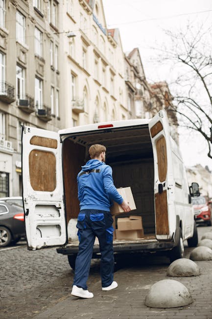 A man wearing a blue jacket, blue trousers, and white sneakers is standing at the rear of a white cargo van parked on a cobblestone street in Notting Hill. He is holding a small cardboard box and appears to be in the process of loading it onto or unloading it from the van, which has its rear doors open revealing an interior lined with wooden panels. Inside the van, there are several other cardboard boxes of various sizes, some placed on the floor. The surrounding environment includes residential buildings with ornate facades, a few parked cars, and leafless trees, indicating an urban setting typical for house removals or small-scale home relocations. The scene captures the logistics involved in furniture transport, packing, and moving processes undertaken by [COMPANY_NAME] as part of their same-day removals service in Notting Hill.