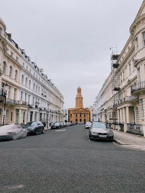 A wide street scene in Notting Hill Gate lined with three- to four-storey white Victorian terraced houses featuring black wrought iron balconies and large windows. Parked cars are visible along both sides of the paved road, with some covered by grey and black car covers. In the foreground, there are metal trolleys and blankets indicating a home relocation or furniture transport process, with a partially visible open moving van or vehicle on the right side near the sidewalk. At the end of the street, a historic building with a clock tower and a distinctive green dome stands prominently under an overcast sky. Scaffolding is visible on one of the buildings on the right, suggesting ongoing maintenance or renovation. This scene captures the logistics and environment typical of house removals in London, with street-level loading and unloading in a residential neighbourhood, natural lighting, and a calm atmosphere.