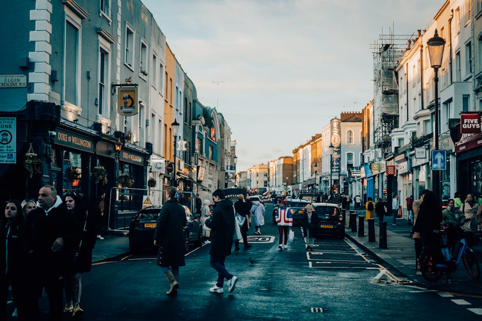 A wide street scene in Notting Hill Gate lined with three- to four-storey white Victorian terraced houses featuring black wrought iron balconies and large windows. Parked cars are visible along both sides of the paved road, with some covered by grey and black car covers. In the foreground, there are metal trolleys and blankets indicating a home relocation or furniture transport process, with a partially visible open moving van or vehicle on the right side near the sidewalk. At the end of the street, a historic building with a clock tower and a distinctive green dome stands prominently under an overcast sky. Scaffolding is visible on one of the buildings on the right, suggesting ongoing maintenance or renovation. This scene captures the logistics and environment typical of house removals in London, with street-level loading and unloading in a residential neighbourhood, natural lighting, and a calm atmosphere.