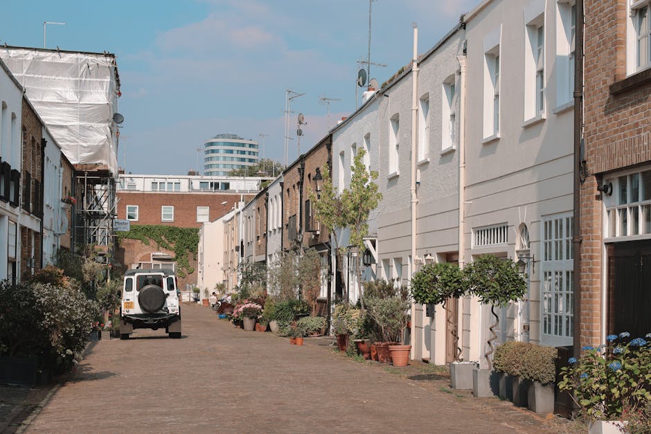 A quiet residential street in Notting Hill with a clear sky and soft natural daylight, featuring a narrow cobblestone pathway lined with closely spaced terraced houses. The houses are built with brick and painted facades, mainly in white or light colours, with small front gardens or potted plants alongside their entrances. Several windows are visible, with some having decorative arches or grilles, and a few houses have external doors open, suggesting ongoing home relocation or packing activities. A white van with a spare wheel mounted on the rear is parked near the centre of the street, likely used for furniture transport and moving logistics. Nearby, a parcel trolley is partially visible, indicating the process of packing or loading household items. Potted plants and small trees are placed along the pavement, adding a touch of greenery. At the far end, a modern multi-storey building is visible against the backdrop of a partly cloudy sky, contrasting with the historic character of the terraced houses. The scene captures a typical moving day or house removal setup within the Notting Hill postcode area, facilitated by Man with Van Notting Hill, illustrating the loading process and the arrangement of residential items for a domestic move.